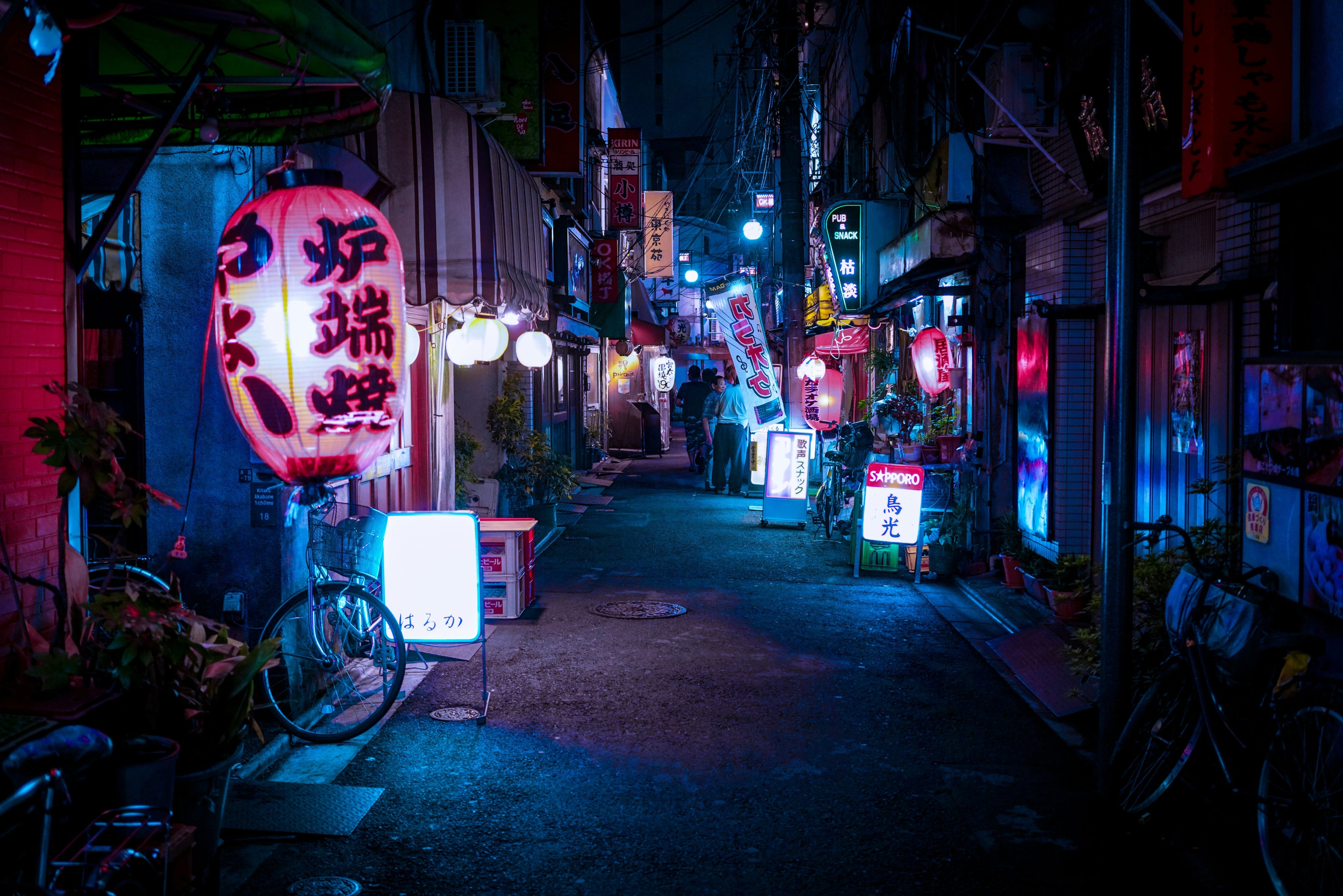 Nachtaufnahme einer schmalen Straße in Tokio, beleuchtet von leuchtenden Laternen und Neonlichtern, mit Fahrrädern und kleinen Geschäften entlang der Gasse.