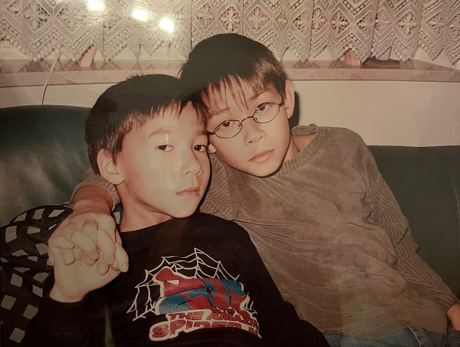 Zwei Jungen sitzen eng nebeneinander auf einem Sofa. Der Junge links trägt ein schwarzes Spider-Man-T-Shirt und schaut ernst, während der Junge rechts eine Brille und ein graues Oberteil trägt. Beide halten sich an den Händen und haben einen nachdenklichen Ausdruck.