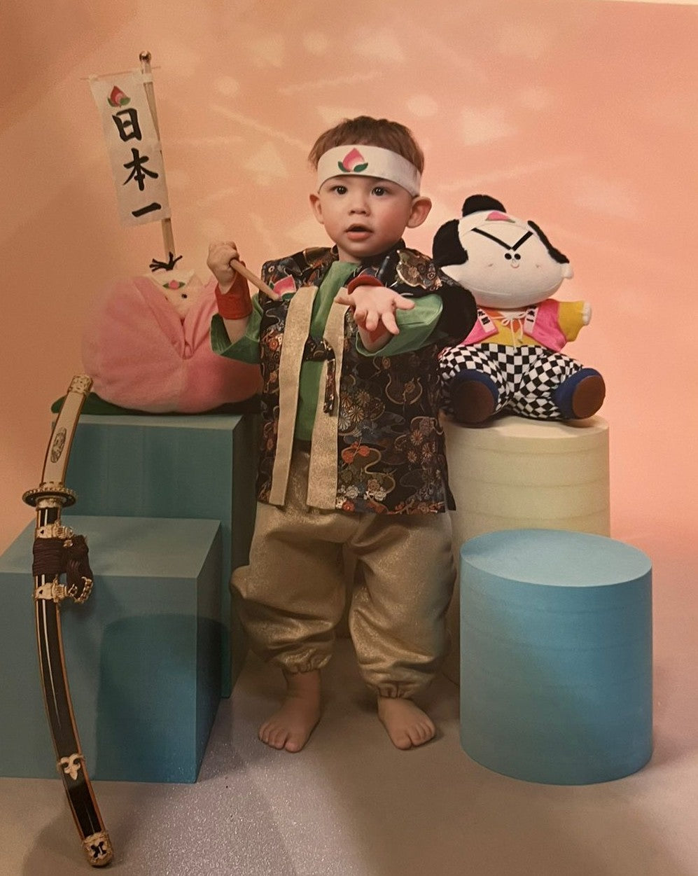 Ein Kleinkind in einem farbenfrohen traditionellen Kostüm, das japanisch inspiriert ist. Es trägt eine Stirnband mit einem Pfirsichsymbol, steht barfuß und hält einen kleinen Stab in der Hand. Neben ihm befinden sich Dekorationen wie ein Schwert, ein Banner mit japanischen Schriftzeichen und eine Stoffpuppe.